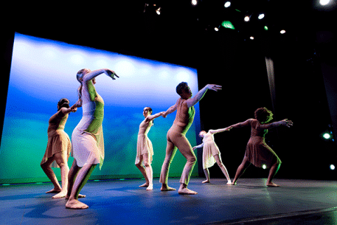 Contemporary dance students perform on a theater stage, extending their arms in coordinated movement against a blue-lit backdrop.