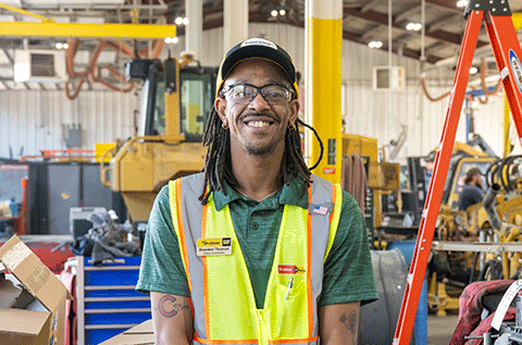 A person wearing safety glasses, a high-visibility safety vest, and a Caterpillar-branded cap stands in a heavy equipment maintenance facility. Construction equipment, a ladder, tools, and workstations are visible throughout the shop in the background.