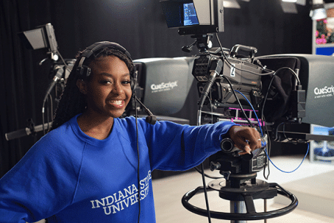 A smiling young woman wearing a headset stands next to a professional video camera in a television studio or media production setting. The camera is mounted on a pedestal and equipped with a teleprompter, with additional studio equipment visible in the background.