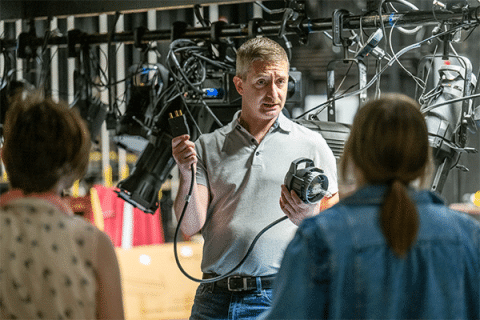 Faculty member demonstrates electrical components and wiring connections to a small group of students in a technical lab filled with cables, equipment, and lighting rigs.