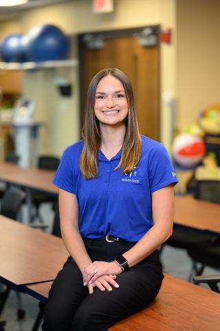 Female with brunette hair wearing a blue Indiana State polo sits on a table in a classroom with therapy equipment, exercise balls, and adaptive toys in the background.