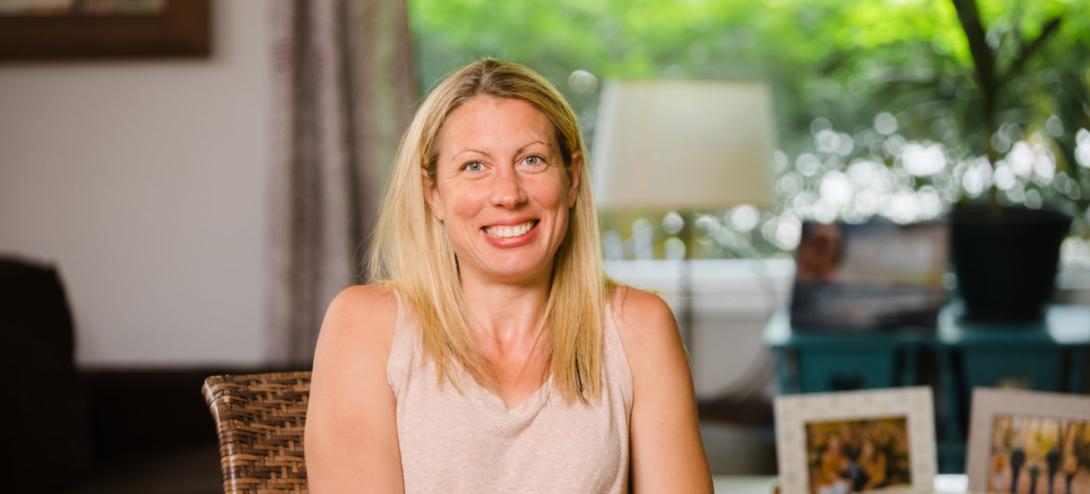 A white female with blonde hair sits indoors. She wears a light beige tank top shirt. In the background, there is a lamp, framed photos, and greenery visible through a window. 