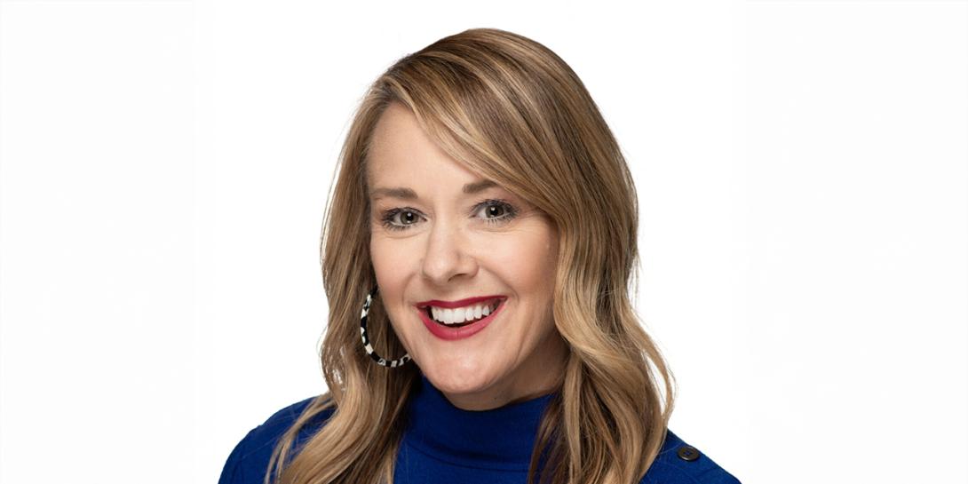 A white female with long, wavy blond hair is wearing a blue dress shirt. The background is plain white. 