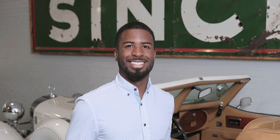 A Black male stands in front of a vintage ca, wearing a white long-sleeved dress shirt. The car is white with a wooden dashboard and tan interior. Behind the person, there is a large green sign with the partially visible word “SINCE” on it. There is a white brick wall visible behind him.