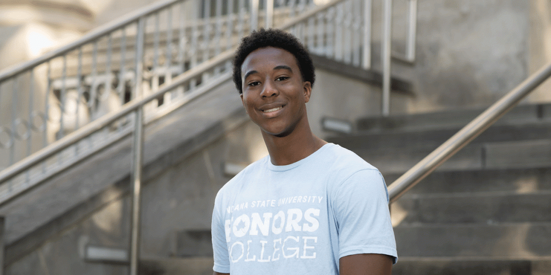 A Black male student is standing in front of a set of outdoor stairs. He is wearing a light blue T-shirt with the text “INDIANA STATE UNIVERSITY HONORS COLLEGE” printed on it in white lettering. The background includes metal railings and stone steps leading up to a building.