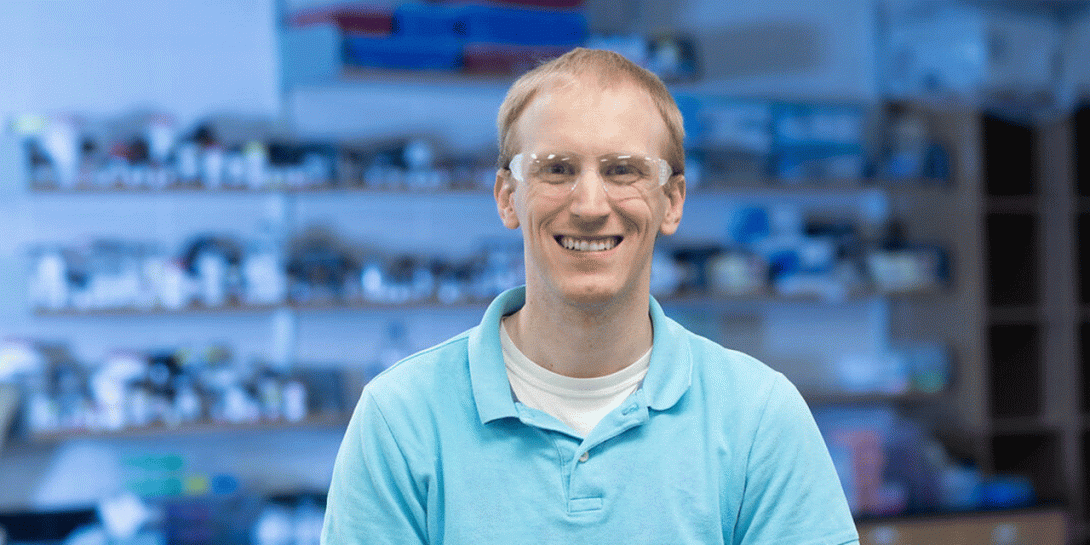 A white male with short blond hair is standing in a laboratory setting, wearing a light blue polo shirt over a white undershirt. Behind him, shelves are filled with various laboratory equipment and supplies, including bottles, containers, and boxes. The background is out of focus. There is a blue tint to the photo.