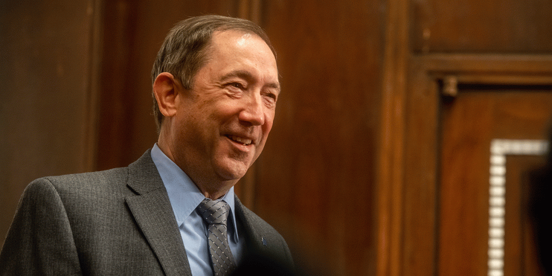 A white male with short brown hair is wearing a grey suit jacket, a light blue dress shirt, and a patterned tie. He stands in front of a wooden paneled wall.